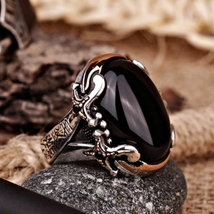 Könnte beinhalten: Ein silberner Ring mit einem schwarzen ovalen Edelstein, der in eine detailreiche silberne Fassung gefasst ist. Der Ring liegt auf einer braunen Oberfläche mit einem Stift und einem Lederarmband.