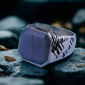 May include: A silver ring featuring a rectangular, pale blue stone. The ring is inscribed with the words "til death". The ring is set against a backdrop of grey rocks, with a blurred background.