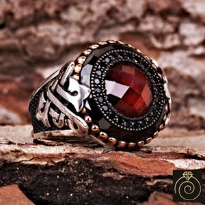 May include: A silver ring featuring a large red gemstone surrounded by smaller black gemstones. The ring has intricate silver detailing and a black onyx band.