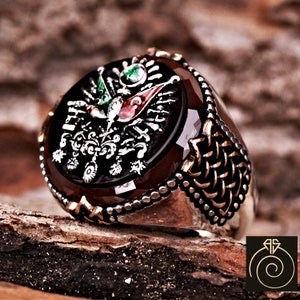 Puede incluir: Un anillo de plata con una piedra preciosa marrón que presenta un diseño detallado de un águila bicéfala con banderas y una corona. La banda del anillo está decorada con un patrón trenzado.