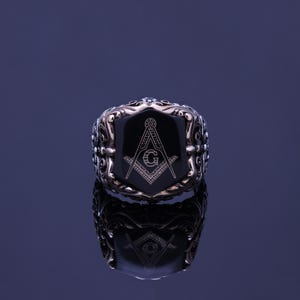May include: A silver ring with a black onyx stone featuring a gold Masonic symbol engraved on the stone.