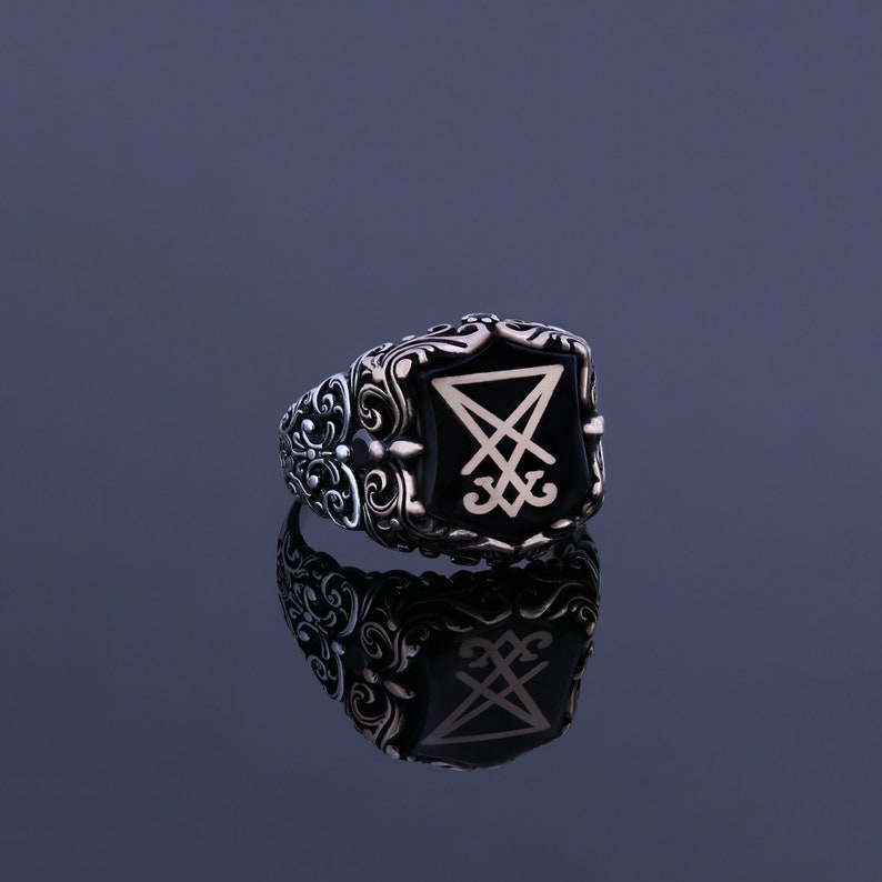 May include: A silver ring with an ornate design and a black stone featuring a white symbol. The ring is reflected in a dark gray surface.
