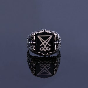 May include: A silver ring with an ornate design featuring a black onyx stone with a white engraved symbol. The symbol is a pentagram with a cross inside.