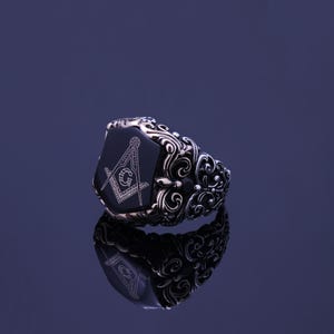 May include: A silver ring with a black onyx stone featuring a detailed engraving of a compass and square, a symbol of Freemasonry. The ring has intricate floral designs on the band.