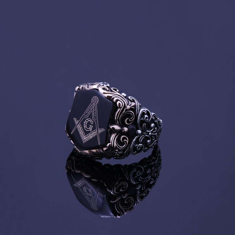 May include: A silver ring with a black onyx stone featuring a detailed engraving of a compass and square, a symbol of Freemasonry. The ring has intricate floral designs on the band.