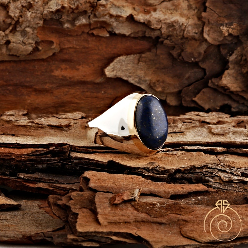 May include: A silver ring with a gold band and a large oval lapis lazuli gemstone. The ring is set against a brown wooden background.