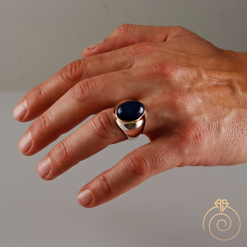 May include: A silver ring with a dark blue oval gemstone set in a gold bezel. The ring is on a person's finger.