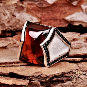 May include: A silver ring with a large, red, faceted gemstone and a white, mother-of-pearl inlay. The ring is decorated with black stones around the edges.