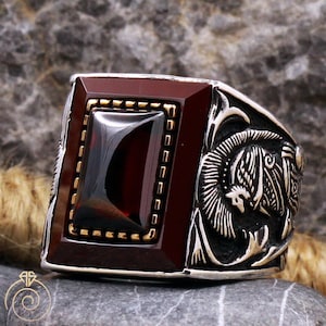May include: A silver ring with a red gemstone set in a gold frame. The ring features an intricate design of a bird with its wings spread.