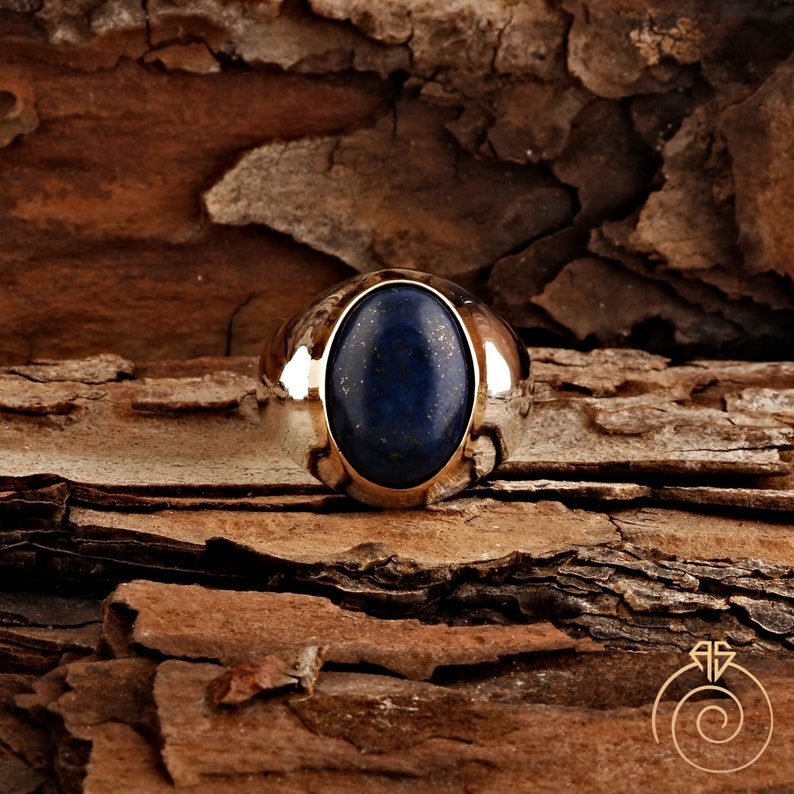 May include: A silver ring with a large oval lapis lazuli gemstone set in a bezel setting. The ring is on a brown wooden surface.