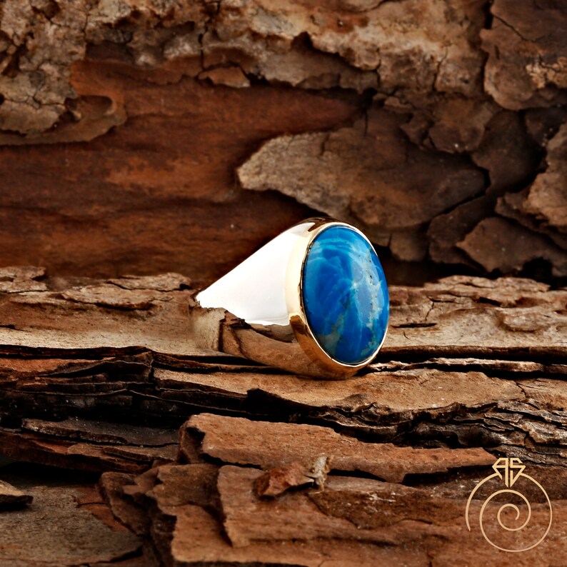 K&ouml;nnte beinhalten: Ein silberner Ring mit einem blauen ovalen Edelstein, der in einer goldenen Fassung sitzt. Der Ring liegt auf einer braunen Holzoberfl&auml;che.