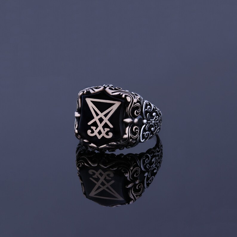 May include: A silver ring with a black onyx stone featuring a white sigil. The ring has an ornate design with intricate details.
