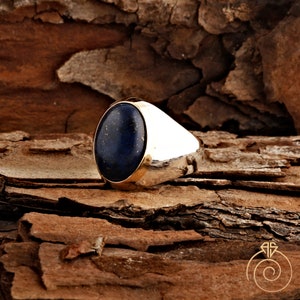 May include: A silver ring with a large oval-shaped blue lapis lazuli gemstone set in a gold bezel. The ring is on a brown wooden surface.