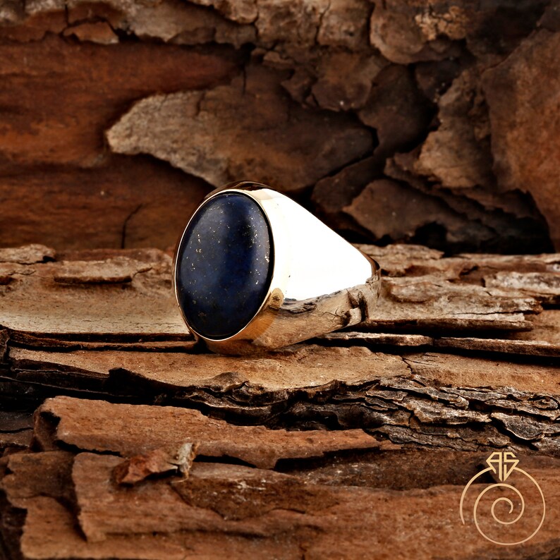 May include: A silver ring with a large oval-shaped blue lapis lazuli gemstone set in a gold bezel. The ring is on a brown wooden surface.