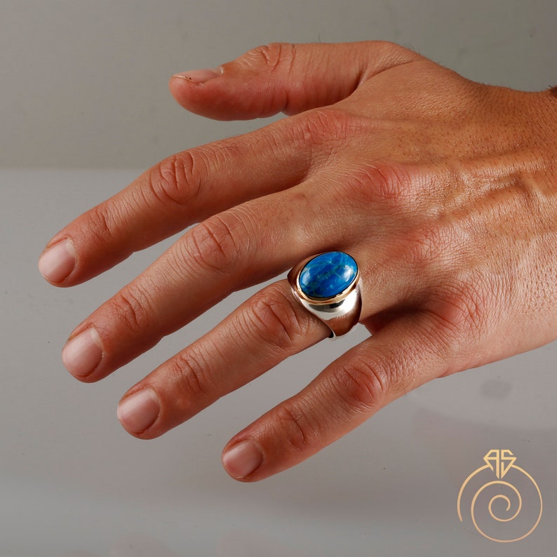 K&ouml;nnte beinhalten: Ein silberner Ring mit einem blauen ovalen Edelstein, der in einer goldenen Fassung sitzt. Der Ring hat ein breites Band und eine polierte Oberfl&auml;che.