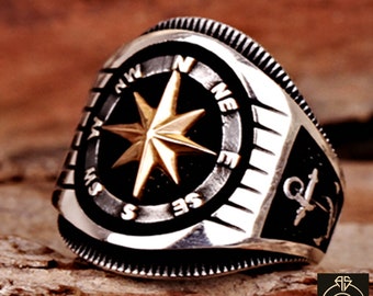 May include: A silver ring with a compass design featuring a gold star and the letters N, E, S, and W. The ring also has an anchor design on the side.