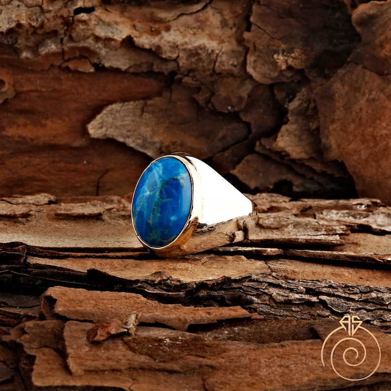 K&ouml;nnte beinhalten: Ein silberner Ring mit einem blauen ovalen Edelstein, der in einer goldenen Fassung sitzt. Der Ring liegt auf einer braunen Holzoberfl&auml;che.