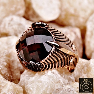 Puede incluir: Un anillo con una gran piedra preciosa de color rojo oscuro engastada en una banda de color dorado. La banda está diseñada de forma intrincada con un patrón texturizado.