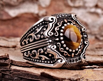 May include: A silver ring with an oval, tiger's eye stone. The ring has an ornate design with scrollwork and small black accents. The ring is set against a textured, brown background.