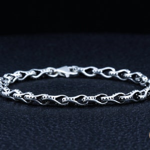 May include: A silver chain bracelet with a detailed, intricate design. The chain is made of small, linked loops with a textured surface.