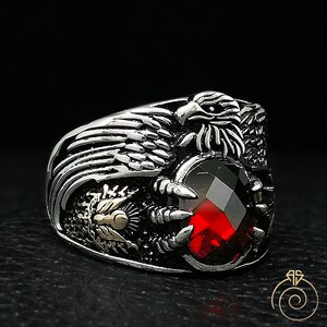 May include: A silver ring with a detailed eagle design holding a red gemstone. The eagle's wings are spread out and the gemstone is set in a claw-like setting.