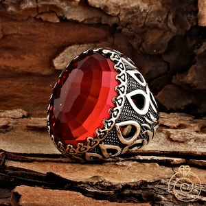 Op de afbeelding: Een zilveren ring met een sierlijk ontwerp en een grote, rode edelsteen.