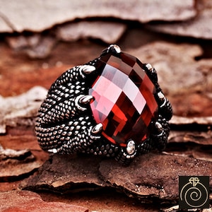 Puede incluir: Un anillo de plata con una gran piedra preciosa roja engastada en una garra. La banda del anillo es texturizada y tiene un diseño detallado.