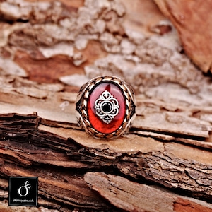 Pu&ograve; includere: Un anello d'argento con una pietra ovale rossa e un design a filigrana d'argento. L'anello &egrave; posto su un pezzo di legno.