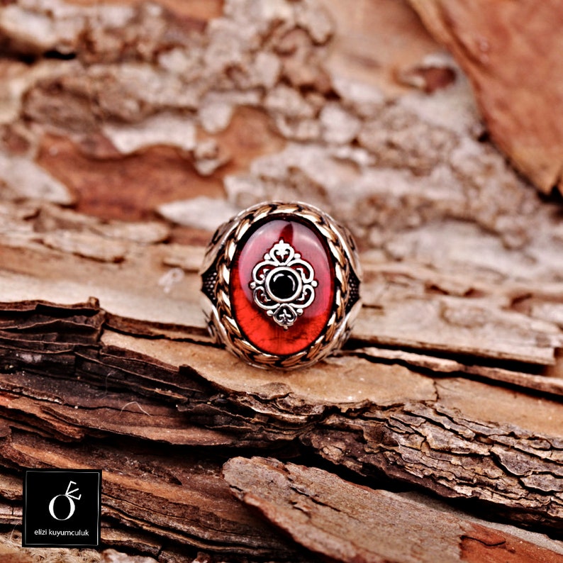 Pu&ograve; includere: Un anello d'argento con una pietra ovale rossa e un design a filigrana d'argento. L'anello &egrave; posto su un pezzo di legno.