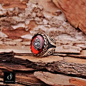 Pu&ograve; includere: Un anello d'argento con una grande pietra rossa ovale incastonata in una montatura d'argento dettagliata. L'anello presenta un design intricato con un piccolo pietra nera al centro della pietra preziosa.