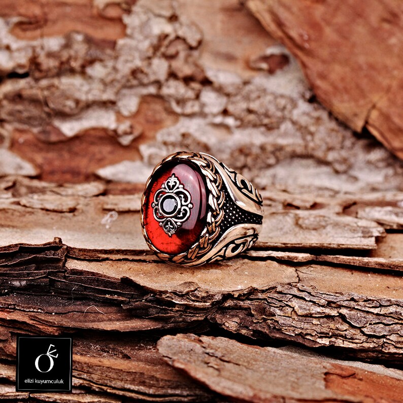 Pu&ograve; includere: Un anello d'argento con una grande pietra rossa ovale incastonata in una montatura d'argento dettagliata. L'anello presenta un design intricato con un piccolo pietra nera al centro della pietra preziosa.