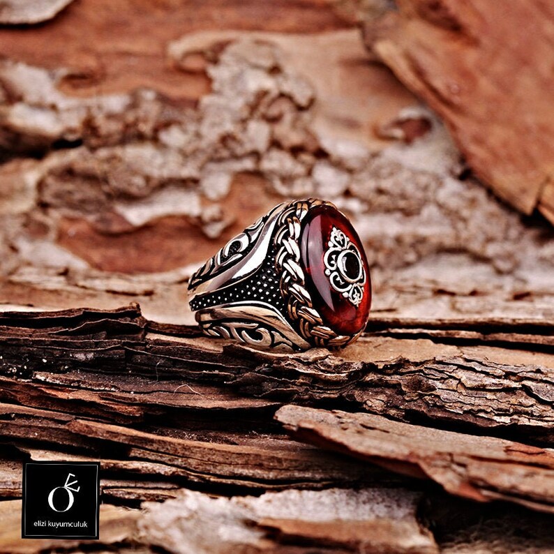 Pu&ograve; includere: Un anello d'argento con una grande pietra rossa ovale incastonata in una montatura d'argento dettagliata. L'anello &egrave; su una superficie di legno.