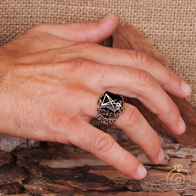 May include: A silver ring with a black onyx stone featuring a gold sigil. The ring has an intricate, ornate design.