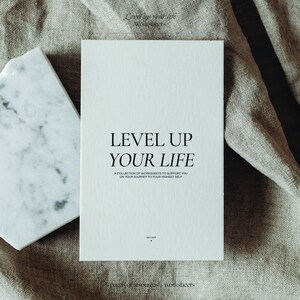May include: A white booklet with the text "Level Up Your Life" in black. The text below reads "A collection of worksheets to support you on your journey to your highest self." The booklet is on a beige fabric surface.