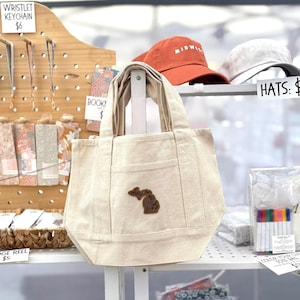 May include: A beige canvas tote bag with a brown leather patch of the state of Michigan. The bag has two handles and a front pocket. In the background are hats and other items for sale.