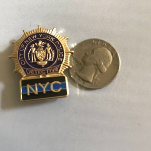 Puede incluir: Pin de metal dorado con el distintivo de detective del Departamento de Policía de la Ciudad de Nueva York con las palabras "City of New York Police" y "Detective" alrededor del distintivo. El pin también tiene una franja azul y negra con las letras "NYC" en blanco.