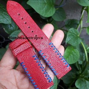 Può includere: Due cinturini per orologio in pelle rossa con cuciture blu. I cinturini hanno una superficie testurizzata e sono mostrati in una mano.