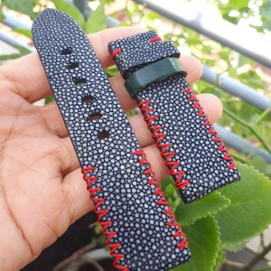 Può includere: Cinturino per orologio in pelle di razza nera con macchie bianche. Il cinturino presenta cuciture rosse e un passante in pelle verde. Il cinturino è mostrato su uno sfondo di foglie verdi.