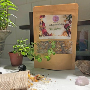 Puede incluir: Una bolsa de papel marrón con una etiqueta que dice "Herbal Bath Salts Skin Soother" y una pequeña bolsa de sales de baño derramadas sobre una superficie blanca. Las sales de baño son blancas con flores y hierbas secas.