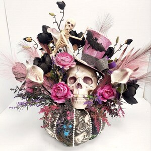 K&ouml;nnte beinhalten: Ein Halloween-Zentrumst&uuml;ck mit einem Sch&auml;del, der einen rosa Zylinder mit einem Fledermaus-Design tr&auml;gt, umgeben von schwarzen und rosa Blumen, einem Skelett und einem dekorativen K&uuml;rbis.