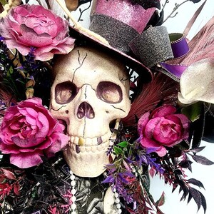 K&ouml;nnte beinhalten: Ein wei&szlig;er Sch&auml;del mit einem goldenen Zahn, der einen rosa und schwarzen Zylinder mit einer schwarzen Fledermaus auf der Oberseite tr&auml;gt. Der Sch&auml;del ist von rosa Rosen, lila Blumen und schwarzen Federn umgeben.