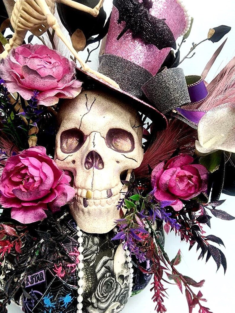 K&ouml;nnte beinhalten: Ein wei&szlig;er Sch&auml;del mit einem goldenen Zahn, der einen rosa und schwarzen Zylinder mit einer schwarzen Fledermaus auf der Oberseite tr&auml;gt. Der Sch&auml;del ist von rosa Rosen, lila Blumen und schwarzen Federn umgeben.