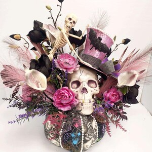K&ouml;nnte beinhalten: Ein Halloween-Zentrumst&uuml;ck mit einem Sch&auml;del, der einen rosa-schwarzen Zylinder mit einem Fledermaus-Design tr&auml;gt. Der Sch&auml;del ist umgeben von rosa und schwarzen Blumen, schwarzen Federn und einer Skelettfigur.