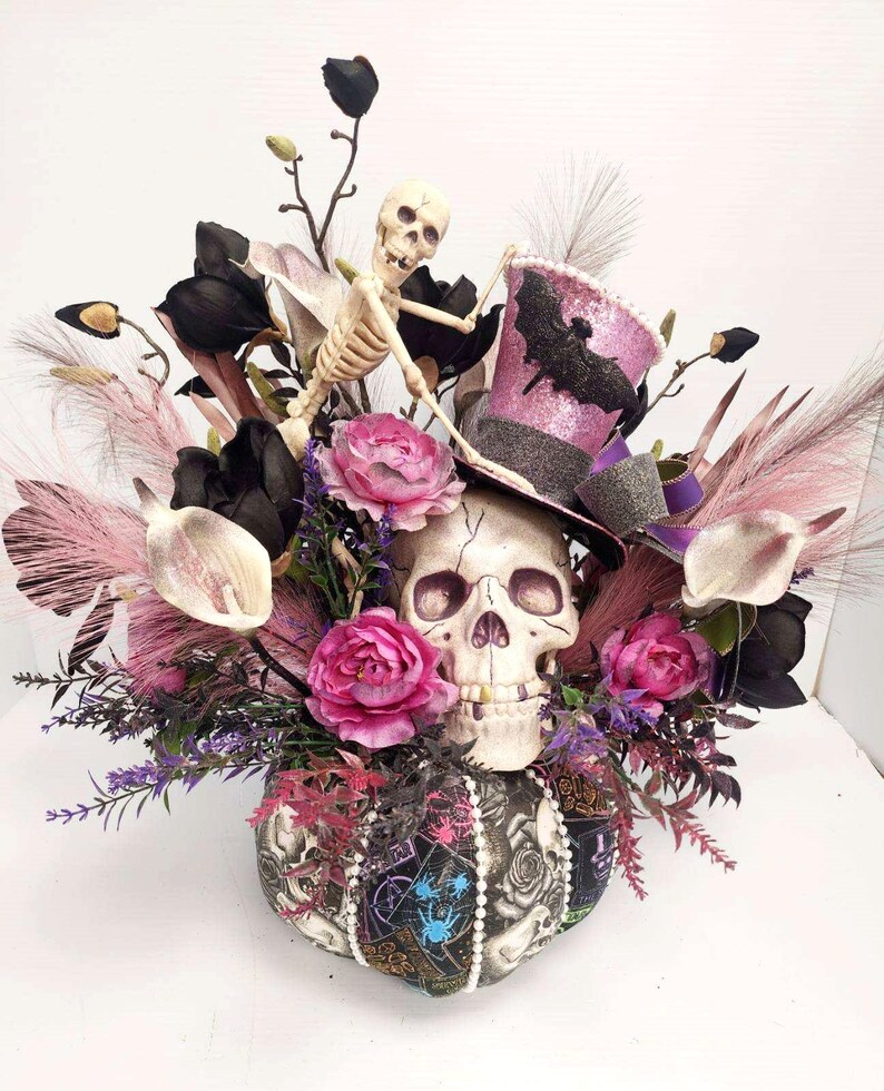 K&ouml;nnte beinhalten: Ein Halloween-Zentrumst&uuml;ck mit einem Sch&auml;del, der einen rosa-schwarzen Zylinder mit einem Fledermaus-Design tr&auml;gt. Der Sch&auml;del ist umgeben von rosa und schwarzen Blumen, schwarzen Federn und einer Skelettfigur.
