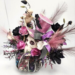 K&ouml;nnte beinhalten: Ein Halloween-Mittelst&uuml;ck mit einem Sch&auml;del, der einen rosa-schwarzen Zylinder tr&auml;gt, umgeben von schwarzen und rosa Blumen und einer Skelettfigur.
