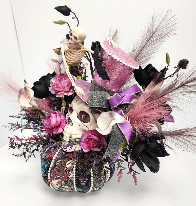 K&ouml;nnte beinhalten: Ein Halloween-Mittelst&uuml;ck mit einem Sch&auml;del, der einen rosa-schwarzen Zylinder tr&auml;gt, umgeben von schwarzen und rosa Blumen und einer Skelettfigur.