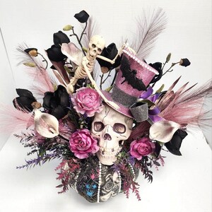 K&ouml;nnte beinhalten: Ein Halloween-Blumenarrangement mit einem Sch&auml;del, der einen rosa und violetten Zylinder mit einem Fledermaus-Design tr&auml;gt. Das Arrangement enth&auml;lt schwarze, rosa und wei&szlig;e Blumen sowie eine Skelettfigur.