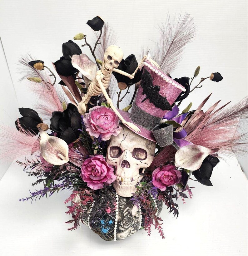 K&ouml;nnte beinhalten: Ein Halloween-Blumenarrangement mit einem Sch&auml;del, der einen rosa und violetten Zylinder mit einem Fledermaus-Design tr&auml;gt. Das Arrangement enth&auml;lt schwarze, rosa und wei&szlig;e Blumen sowie eine Skelettfigur.