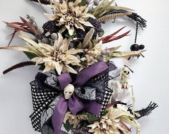 May include: A Halloween-themed wreath with beige dahlias, burgundy leaves, and a black and white patterned bow. A skeleton bride sits on a broom, and a black spider hangs from the wreath. The text "AC Inspired Wreaths" is visible.