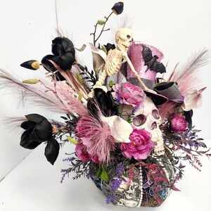 K&ouml;nnte beinhalten: Ein Halloween-Blumenarrangement mit einem Sch&auml;del mit Zylinder, einem Skelett, schwarzen und pinkfarbenen Blumen und verschiedenen Gr&uuml;npflanzen.